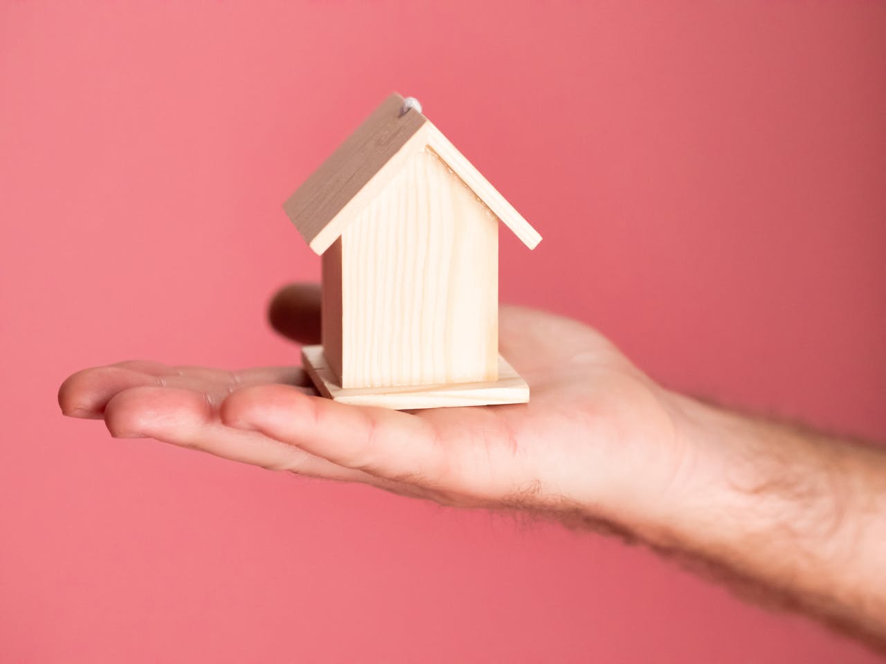 pexels-photo-7579354 Wooden house model in hand symbolizing real estate concepts on pink background.